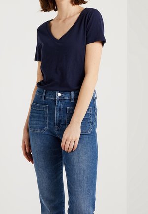 Kvinde iført en marineblå V-hals kortærmet skjorte, stoppet ned i højttaljede blå jeans med forlommer.