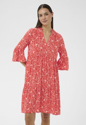 Donna con capelli lisci scuri che indossa un vestito rosso al ginocchio con stampa floreale bianca e maniche a campana a tre quarti, in piedi contro uno sfondo bianco.