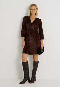 Mujer vestida con un vestido de leopardo marrón hasta la rodilla y mangas de 3/4, sosteniendo un bolso de mano negro y botas de caña alta marrón oscuro.