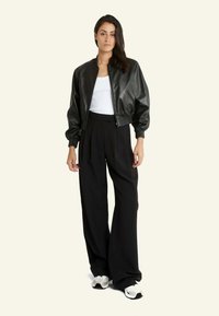 Veste bomber en simili cuir noir avec des poignets côtelés, associée à un pantalon noir ample et des baskets blanches, le tout sur un fond neutre.