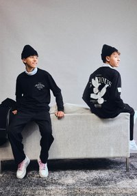 Két fiatal férfi fekete sapkában és fekete grafikás pulóverben, háttal ülve egy bézs kanapén egy minimalista szobában.
