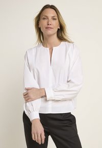 Weiße Bluse mit langen Ärmeln, hochgeschlossen und lässig geschnitten, mit Puffärmeln und geknöpfte Manschetten; glatte Stofftextur.