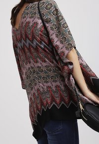 Flerfärgad mönstrad poncho med V-hals, prydd med zigzag-mönster i rött och grönt, samt en svart nederdel. Matchad med en svart axelväska.