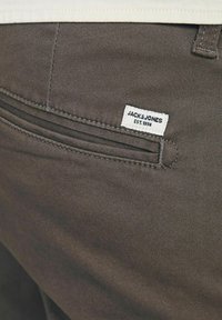 Braune Stoffhose mit einem Reißverschlusstaschen-Detail. Ein weißes Etikett mit "JACK & JONES EST. 1990" ist in der Nähe der Taschennähte genäht. Glatte Textur.