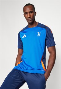 Camisola atlético azul com detalhes laranja, apresentando mangas curtas e decote redondo. O material parece ser de absorção de humidade, exibindo logótipos brancos.