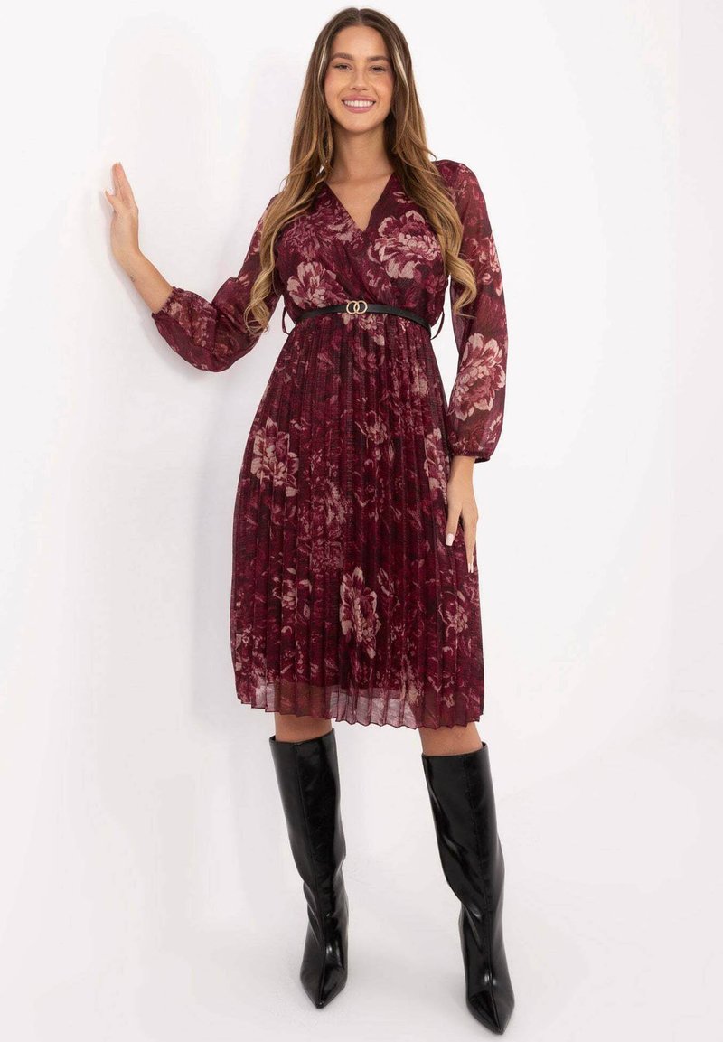 Dunkelrotes, floral gemustertes, plissiertes Kleid mit langen, transparenten Ärmeln, einem V-Ausschnitt und einem schwarzen Gürtel mit doppelter Metall-Schnalle. Kombiniert mit knielangen schwarzen Stiefeln.