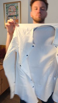 Camisa branca de botões levantada, com botões azuis, colarinho arredondado e uma textura suave. O fundo inclui um armário desfocado e uma obra de arte.