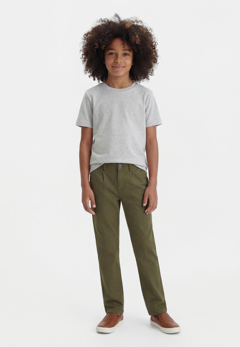 Enfant debout, mains détendues, portant un t-shirt gris clair, un pantalon vert olive et des chaussures marron sans lacets sur un fond blanc uni.