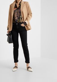 Blazer color camel sobre una camisa negra con estampado, combinado con jeans negros remangados y zapatos planos puntiagudos con una correa negra. Bolso negro.