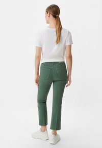 Zielone przednie spodnie z dwiema tylnymi kieszeniami, zestawione z białą koszulką. Obuwie to białe sneakersy. Tło jest jednolite białe.