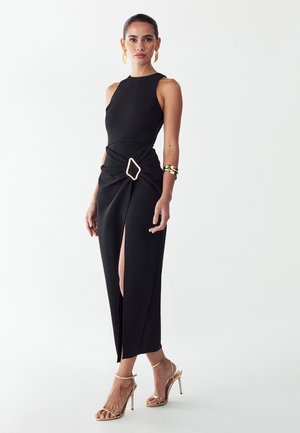 Kobieta ubrana w czarną, bezrękawową sukienkę midi z bocznym rozcięciem i złotą klamrą, złote sandały na wysokim obcasie, złote kolczyki i bransoletkę.