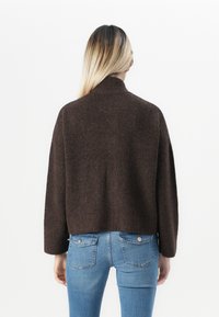 Personne avec de longs cheveux blonds portant un pull marron foncé et un jean bleu, debout de dos contre un fond clair.