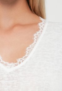 Hvid bluse med V-udskæring og delikat spidsdesign, lavet af letvægts, tekstureret stof. Nærbillede af detaljen omkring udskæringen.