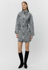 Grå denimklänning med hög krage, långa ärmar och framfickor. Har dragkedjestängning och insydd midja, kombinerad med knähöga svarta stövlar.