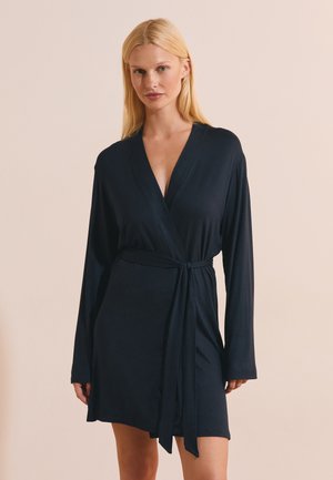 Frau mit langen blonden Haaren, die ein dunkelblaues Wickelkleid mit langen Ärmeln trägt, das an der Taille gebunden ist, steht vor einem beigen Hintergrund.