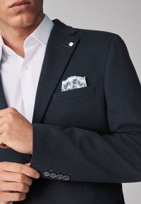 Blazer texturizado en color marino con solapa de muesca, dos botones y emblema bordado. El pañuelo de bolsillo presenta un patrón geométrico en azul claro y blanco.