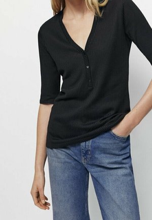 Model trägt ein schwarzes, geripptes V-Ausschnitt-Oberteil mit einer halben Knopfleiste, kombiniert mit hellblauen Jeans, was einen lässigen Stil zeigt.