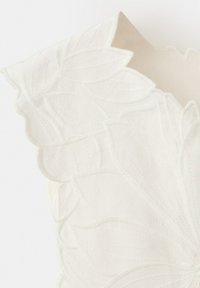 Pièce de tissu écru avec des bords festonnés et un motif brodé texturé en forme de feuille sur un fond lisse.