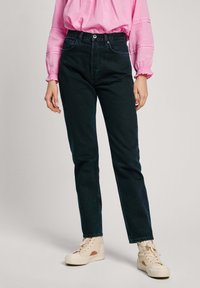 Vaqueros de mezclilla oscura con cintura alta, diseño de pierna recta y detalles de costura sutiles, combinados con una blusa rosa y zapatillas de color claro.