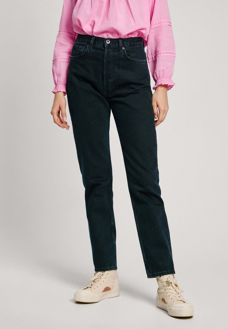 Vaqueros de mezclilla oscura con cintura alta, diseño de pierna recta y detalles de costura sutiles, combinados con una blusa rosa y zapatillas de color claro.