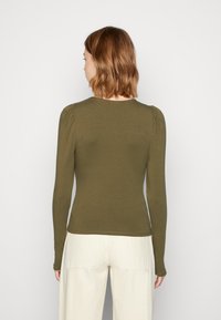 Frau mit schulterlangen braunen Haaren, die ein olivgrünes Langarmoberteil und off-white Hosen trägt, von hinten vor einem einfarbigen Hintergrund betrachtet.