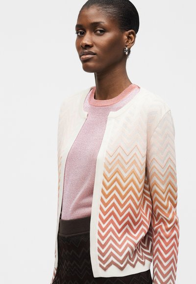 Γυναίκα με κοντά μαλλιά φορά ασημένια σκουλαρίκια κρίκους, ροζ μπλούζα και λευκή ζακέτα με πορτοκαλί και καφέ ζιγκ-ζαγκ σχέδια στα μανίκια.
