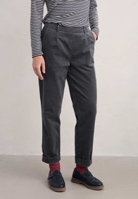 Pilkos, aukštai juosmens kelnės, pagamintos iš tekstūruoto audinio, su klostėmis, šoninėmis kišenėmis ir sukamuoju kraštu, derinamos su tamsiais batais ir raudonomis kojinėmis.