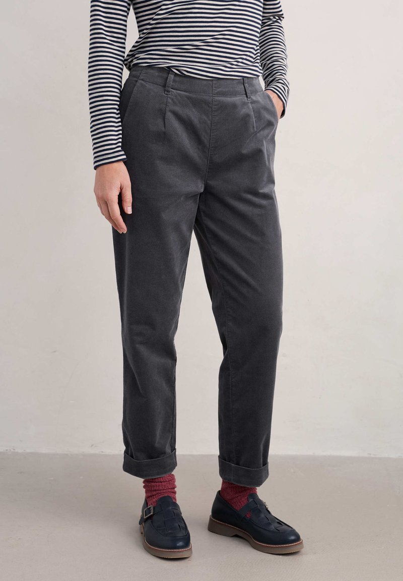 Pilkos, aukštai juosmens kelnės, pagamintos iš tekstūruoto audinio, su klostėmis, šoninėmis kišenėmis ir sukamuoju kraštu, derinamos su tamsiais batais ir raudonomis kojinėmis.