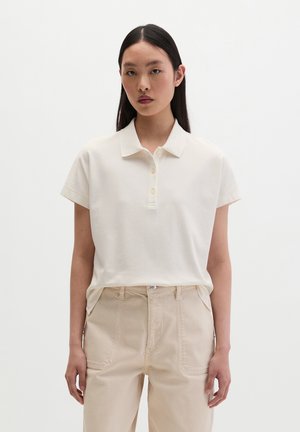 Kobieta o długich ciemnych włosach nosi białą koszulkę polo z krótkim rękawem i beżowe spodnie z wysokim stanem na jednolitym białym tle.