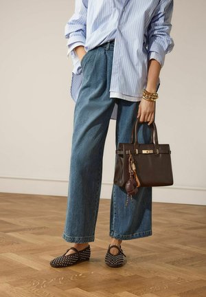 Vrouw draagt losse spijkerbroek, gestreept shirt, gespenste platte schoenen, houdt een donkere leren handtas met decoratieve bedels vast in een neutrale binnenomgeving.