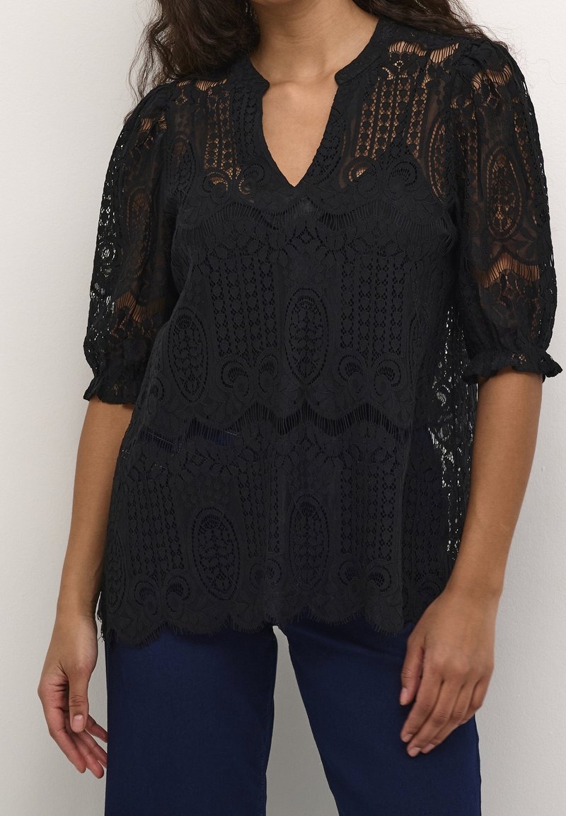 Femme portant une blouse en dentelle noire avec un col en V et des manches bouffantes mi-longues, associée à un pantalon bleu foncé sur un fond uni.