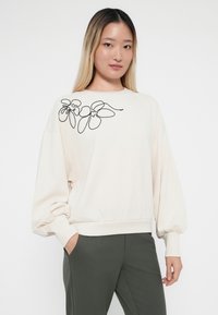 Crème sweatshirt met ballonmouwen, geribbelde manchetten en zoom. Heeft zwarte bloemige borduursels op de linker schouder. Gecombineerd met donkergroene broek.