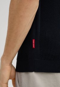 Marineblauwe gebreide trui met een gestructureerd patroon, een geribbelde zoom en een rode label aan de zijkant. Close-up toont de arm en stofdetails.