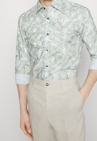 Lichtgewicht, langarm overhemd in het wit met een groen bladerenpatroon, voorzien van groene knopen. Gecombineerd met beige linnen broeken met een soepele textuur.