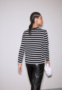 Triibuline must-valge pika varrukaga top, mida on kombineeritud mustade nahksete pükstega. Modell hoiab käes valget tekstuurse mustriga õlakotti ja tal on punutud soeng.