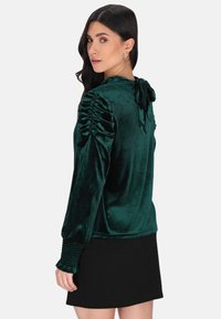 Grüne Samtbluse mit Raffung an den Ärmeln und einem Bindeband auf der Rückseite. Die langen Ärmel haben geraffte Bündchen. Kombiniert mit einem schwarzen Rock.
