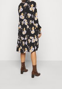 Vrouw in een zwart bloemen midi-jurk, gecombineerd met bruine enkellaarsjes met hak, staand met de rug naar de camera tegen een effen achtergrond.