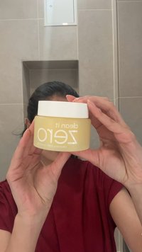 Osoba trzymająca okrągły słoik balsamu oczyszczającego "Clean It Zero" z żółtym opakowaniem i białą nakrętką w łazience z neutralnymi płytkami.