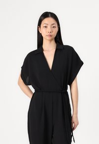Svart jumpsuit med korte ermer, V-hals og flettet belte. Laget av lett materiale, med en avslappet passform og glatt tekstur.
