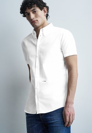 Mladý muž s kučeravými vlasmi, oblečený v bielej krátkorukávovej košeli na gombíky a modrých džínsoch, stojaci pred svetlosivým pozadím.