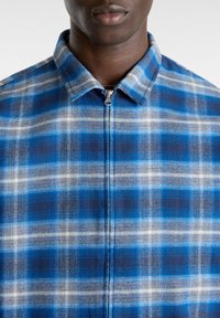 Sininen ja harmaa ruudullinen takki, jossa on vetoketju, kaulus ja tekstuuri kangas. Kuosi koostuu vaihtelevista väreistä olevista neliöistä.