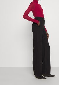 Maglietta rossa a maniche lunghe con collo alto; abbinata a pantaloni neri a gamba larga con tasche frontali e dettagli con bottoni.