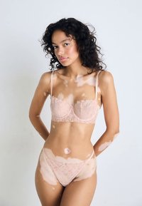 Spitzen-Bralette und passender Tanga in einem sanften Nude-Ton, mit floralen Mustern, verstellbaren Trägern und einer feinen Verzierung. Glatte Textur.
