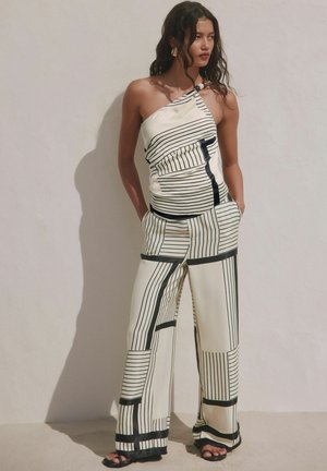 Kobieta o długich ciemnych włosach, ubrana w biały kombinezon z jednym ramieniem i czarnymi geometrycznymi paskami, z rękami w kieszeniach, stojąca na tle gładkiej ściany.