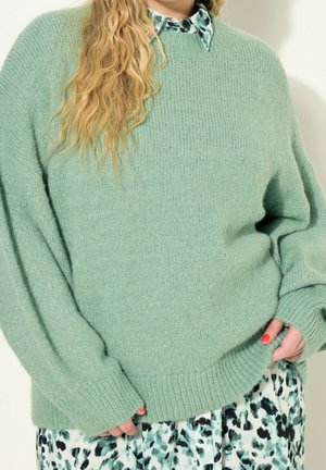 Woman wearing a mint green knit sweater over a black-and-white patterned button-up dress, holding the sweater hem with painted nails.