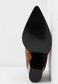 Brun ruskind højhælet sko med spids tå og en blank sort sål, med en formet hæl og glat tekstur.