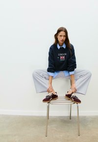 Felpa navy con design "LONDON" e bandiera del Regno Unito, camicia colletto azzurro chiaro, pantaloni grigi a gamba larga, sneakers bordeaux con lacci testurizzati.