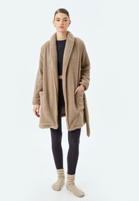 Accappatoio beige, soffice, a maniche lunghe con colletto a scialle, due tasche frontali e una cintura annodata, indossato sopra leggings scuri e calze di maglia.
