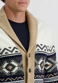 Gebreide cardigan in crème, marineblauw en grijs met geometrische patronen, bruine knoopsluiting en een brede beige kraag. Gestructureerde stof met verschillende accenten.