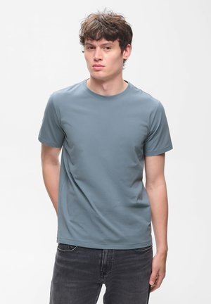 Jeune homme aux cheveux bruns courts et bouclés, portant un t-shirt bleu uni à manches courtes et un jean foncé, debout devant un fond blanc.